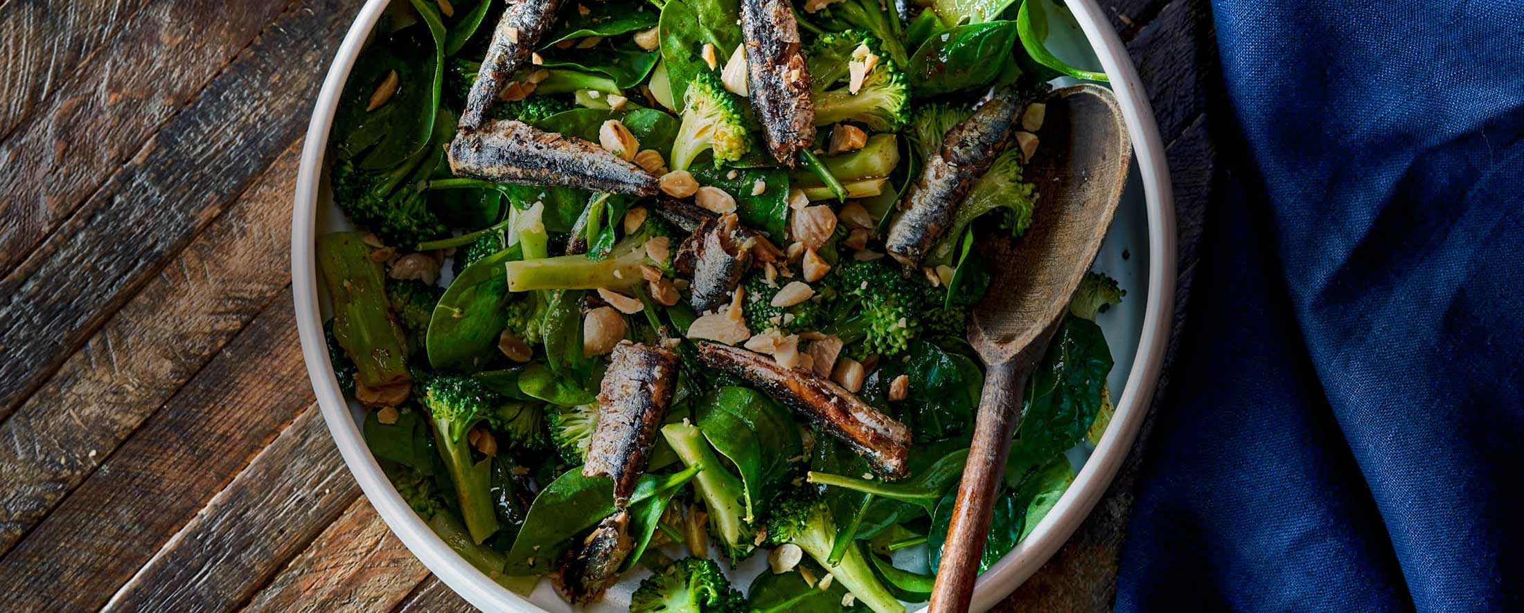 A bowl of dark green salad topped with Patagonia Provisions tinned anchovies, nuts, and broccoli